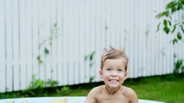 Um menino de quatro anos encontra-se em uma piscina inflável das crianças, salpicando a água, sorrindo. Brinquedos flutuantes nas proximidades. No jardim, no verão. Ele diverte-se . — Vídeo de Stock
