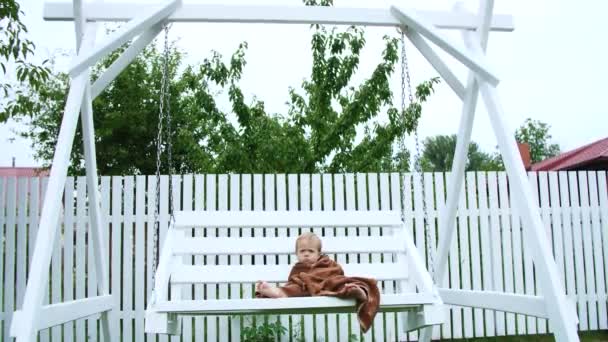 Una niña de un año, envuelta en una toalla, con una cara sucia, sola sentada en un columpio en el jardín, en el verano. Tiene una mirada triste. Ella quiere dormir. — Vídeos de Stock