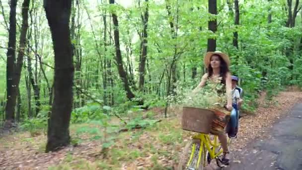 Eine Frau mit Hut und Kleid, mit einem Korb voller Blumen, zusammen mit einem kleinen Kind, Fahrrad fahren, im Wald, im Sommer. das Kind sitzt in einem speziellen Stuhl — Stockvideo