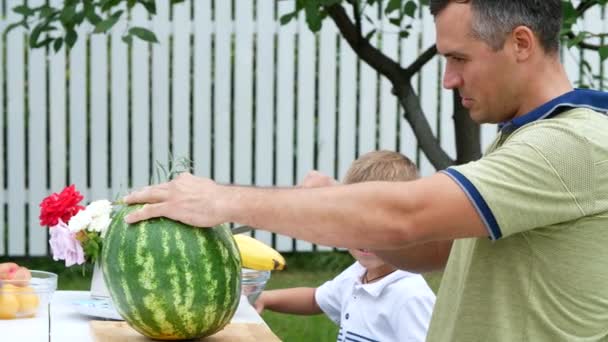 Een jong gezin met twee kleine grappige kinderen zitten op een tafel in de tuin, in de zomer. Papa snijdt een grote watermeloen, gaat voor de behandeling van zijn familie. lunch met de familie. — Stockvideo