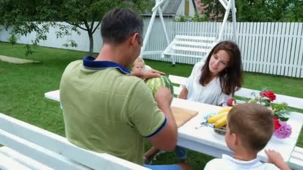 Egy fiatal család két kis vicces gyerek ül egy asztalnál, a kertben, a nyári időszakban. Apa egy nagy görögdinnye darabok, kezelésére a családja lesz. ebéd, a család. — Stock videók