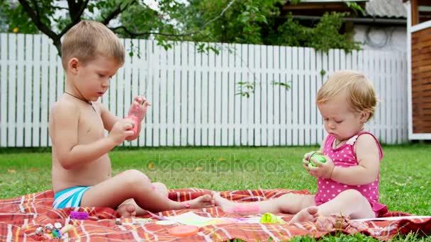 Niños pequeños, un niño de cuatro años y una niña de un año, hermano y hermana, juegan juntos, pintan con pinturas de dedos, en el jardín, sentados en una manta, en la hierba, el césped, en verano — Vídeos de Stock