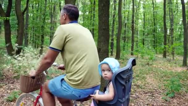 Ein Mann mit einem Kind auf einem Fahrrad im Wald, im Sommer sitzt das Kind in einem speziellen Stuhl — Stockvideo