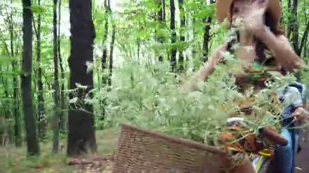 Kobieta w kapeluszu i sukienka, z koszem kwiatów, wraz z małym dzieckiem, jeździć rowerze, w lesie, latem. dziecko siedzi w fotelu na specjalne — Wideo stockowe