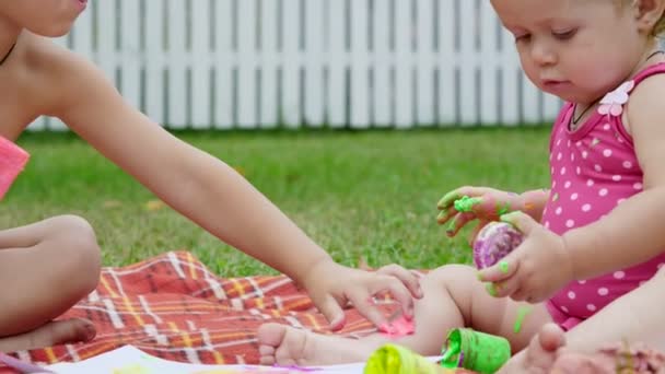 Niños pequeños, un niño de cuatro años y una niña de un año, hermano y hermana, juegan juntos, pintan con pinturas de dedos, en el jardín, sentados en una manta, en la hierba, el césped, en verano — Vídeo de stock