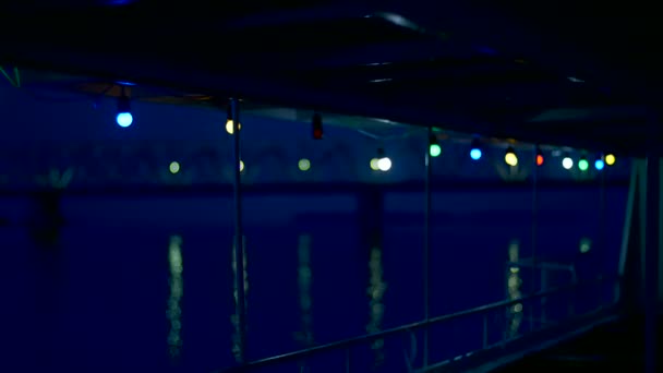 Natt til kveld, langs elven, flyter fergen nær den store broen. Dekket er dekorert med fargede lys. Lysene reflekteres i vannet. utsyn fra dekket . – stockvideo
