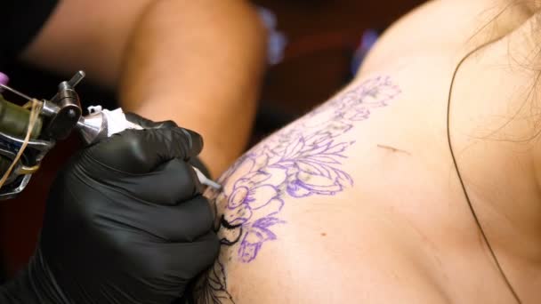 Primer plano, en un salón de tatuajes, un especialista está haciendo un tatuaje en la espalda de las mujeres, adorno floral de pintura negra. un hombre trabaja con guantes negros especiales, en equipo especial . — Vídeo de stock