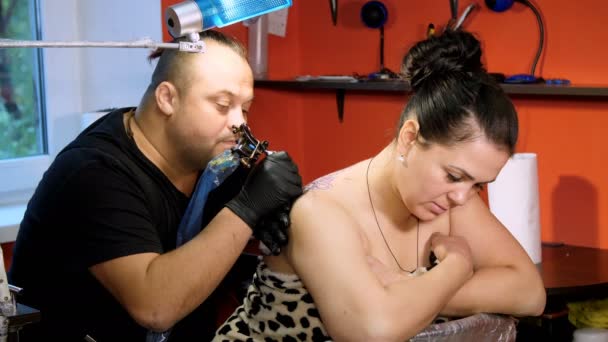 En un salón de tatuajes, un especialista está haciendo un tatuaje en la espalda de las mujeres, un adorno floral. un hombre trabaja con guantes especiales, con equipos especiales. dibujo se hace con pintura negra . — Vídeos de Stock