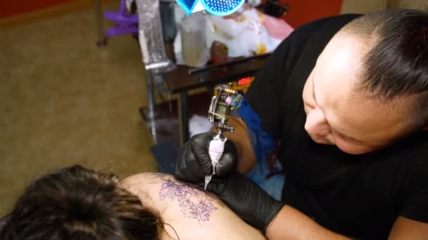 En un salón de tatuajes, un especialista está haciendo un tatuaje en la espalda de las mujeres, un adorno floral. un hombre trabaja con guantes especiales, con equipos especiales. dibujo se hace con pintura negra . — Vídeo de stock