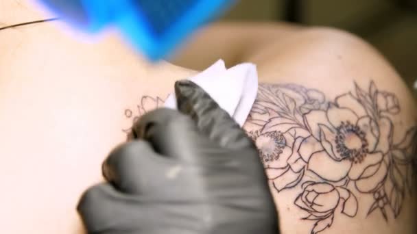 Närbild, i en tatuering salongen, en specialist gör en tatuering på kvinnans rygg, svart färg blommor prydnad. en man fungerar i speciella svarta handskar, på särskild utrustning. — Stockvideo