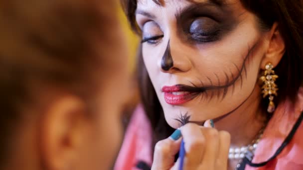 Halloweenfeest, Visagist trekt een verschrikkelijke make-up op het gezicht van een brunette vrouw voor een Halloween party. op de achtergrond, wordt het landschap in de stijl van Halloween gezien — Stockvideo