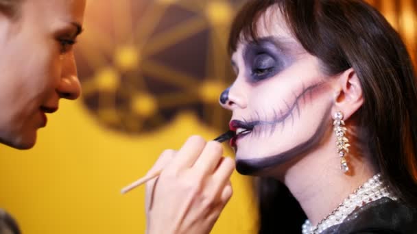 Halloweenfeest, Visagist trekt een verschrikkelijke make-up op het gezicht van een brunette vrouw voor een Halloween party. op de achtergrond, wordt het landschap in de stijl van Halloween gezien — Stockvideo