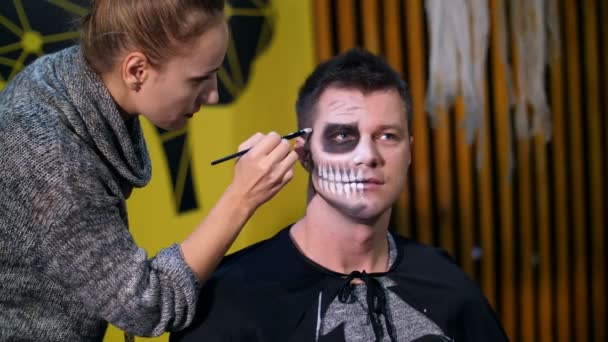 Halloweenfeest, Visagist trekt een verschrikkelijke make-up op het gezicht van een man voor een Halloween party. op de achtergrond, wordt het landschap in de stijl van Halloween gezien — Stockvideo