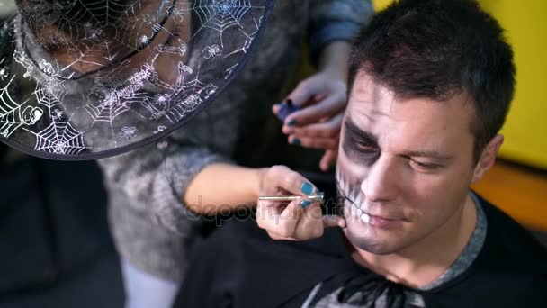 Halloween party, detail, make-up artist nakreslí hrozné make-up na tváři muže na Halloween party. v pozadí je vidět krajina ve stylu Halloween — Stock video