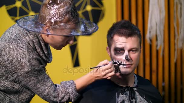 Halloween-Party, Maskenbildner zeichnet ein schreckliches Make-up auf das Gesicht eines Mannes für eine Halloween-Party. im Hintergrund ist die Szenerie im Stil von Halloween zu sehen — Stockvideo