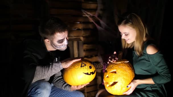Date in style of Halloween party, night, twilight, in the rays of light, guy with a girl dressed in costumes and with a terrible makeup are holding two large Halloween pumpkins — Stock Video
