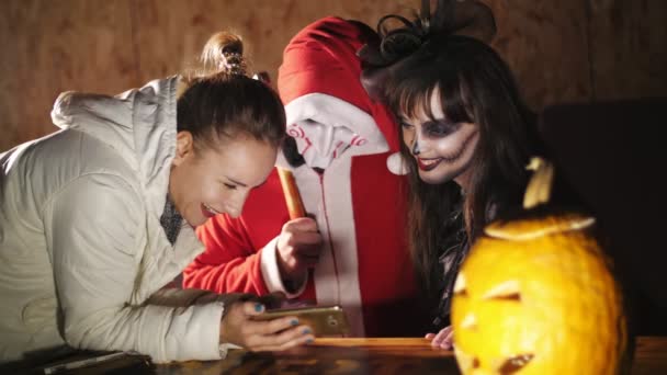Halloween party, night, twilight, in the rays of light, a guy dressed as an evil santa and a woman with a terrible makeup and their friend girl are looking into the phone, reading smth — Stock Video