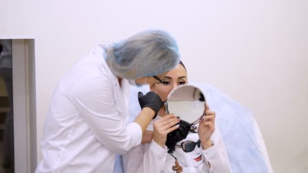 Clínica, sala de cosmetología, hermosa mujer mirando en el espejo, el médico discute el procedimiento de corrección de la forma de los labios con ácido hialurónico , — Vídeos de Stock