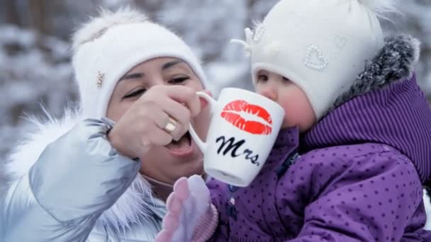 Mot bakgrund av skogen vinter, en ung kvinna, ger mamma en drink till en ett-årig dotter från en kopp. de är klädda i roliga hattar med bumbon. — Stockvideo