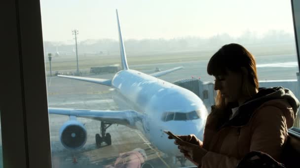 A repülőtéren, a váróban a háttérben egy ablak kilátással a repülőgépek és a kifutópálya áll egy fiatal nő, írja a telefon. Lásd őt sziluett — Stock videók