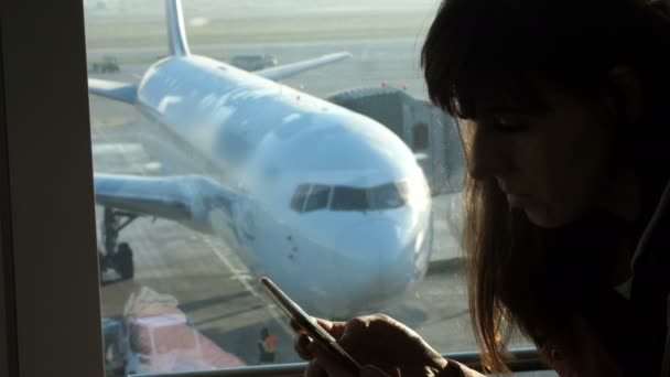 控え室で、空港でウィンドウの背景に飛行機や、滑走路を見下ろす座っている若い女性が携帯電話で入力します。彼女のシルエットを参照してください。 — ストック動画