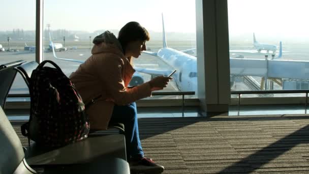 En el aeropuerto, en la sala de espera, sobre el fondo de una ventana con vistas a los aviones y la pista, se sienta una joven mujer escribiendo en el teléfono. ver su silueta — Vídeos de Stock