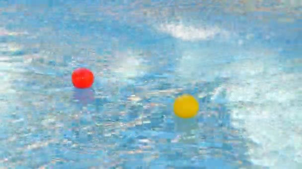Крупный план. В бассейне плавают два маленьких цветных шарика. сверху брызгает водой из фонтана. Красочные резиновые шарики в воде — стоковое видео
