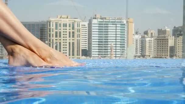 Close-up de belas pernas femininas, pés, na piscina superior do telhado com bela vista para a cidade, contra um pano de fundo de arranha-céus da cidade. Pés femininos em água azul . — Vídeo de Stock