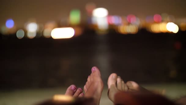 Fiatal párok lábak, láb, két szerelmes a tengerparton repülőtér közelében a víz, éjszaka, fények, a város felhőkarcolók, a ragyog a fény ellen — Stock videók