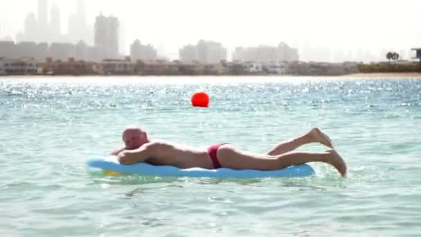 Um homem está descansando em um colchão inflável, nadando no mar, tomando sol. descansar empresário depois de uma semana de trabalho duro. no pano de fundo dos arranha-céus da cidade — Vídeo de Stock