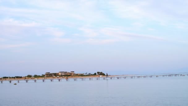 Old, abandoned pier, red sea is calm, windless weather, there are clouds in the sky, you can see a long pier on the horizon. man is walking along the pier — Stockvideo