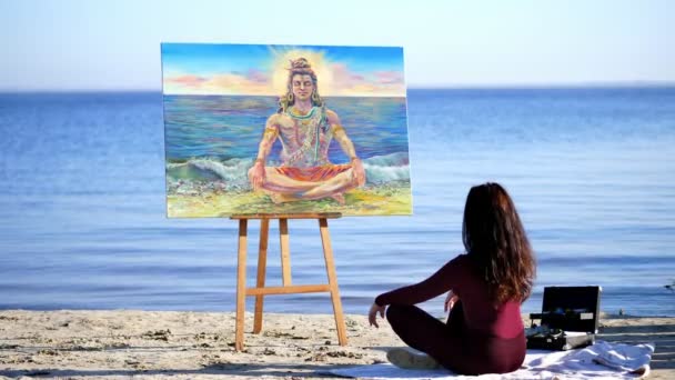 En verano, en la orilla del río, en la playa. al amanecer, una hermosa artista se sienta en una posición de loto frente a un caballete con una imagen de una deidad. meditación . — Vídeos de Stock