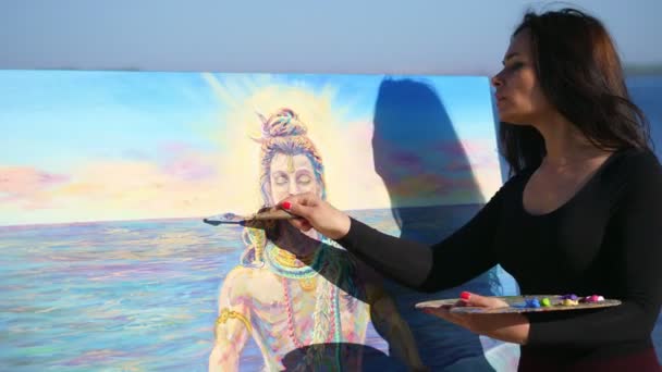 Été, à l'extérieur, près de la rivière, une belle artiste femme dessine, applique des peintures sur toile avec une petite spatule spéciale, en utilisant une technique de dessin spécial. tient une palette avec des peintures — Video
