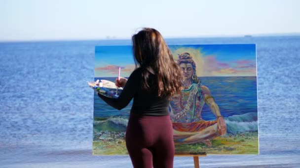 Лето, рядом река, на пляже. На рассвете красивая художница в обтягивающем костюме рисует картину. бог Шива изображен на фотографии — стоковое видео