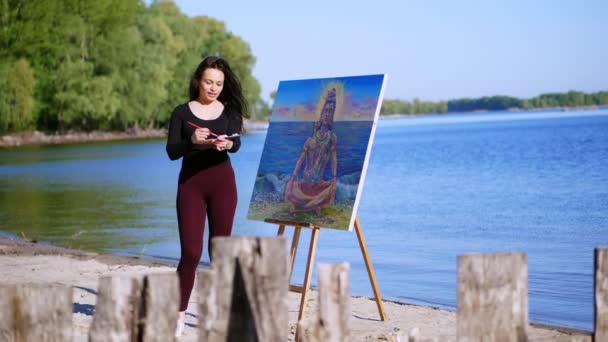 夏には、ビーチでの川の近く。日の出は、タイトなスーツで美しい女性アーティストは、画像を描画します。彼女の髪が風になびきます。シヴァ神が絵に描かれています。 — ストック動画