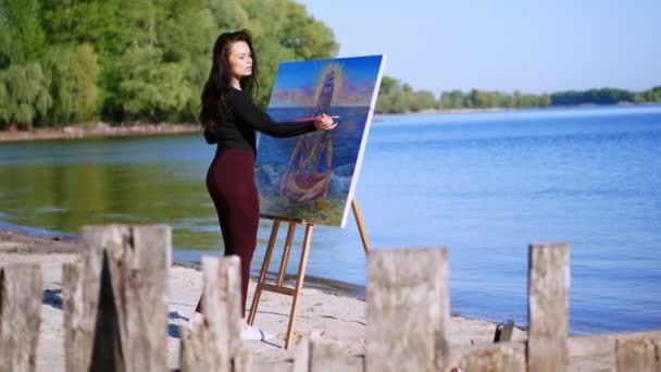 Estate, vicino al fiume, sulla spiaggia. all'alba, una bella artista donna in un abito stretto disegna un disegno. dio Shiva è raffigurato sulla foto — Video Stock