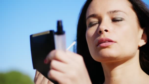 Portret van een mooie veertig jaar oude vrouw, brunette met groene ogen. ze ziet er in kleine spiegel in poeder doos en kleuren lippen met lipgloss. zomer, tegen de blauwe hemel — Stockvideo