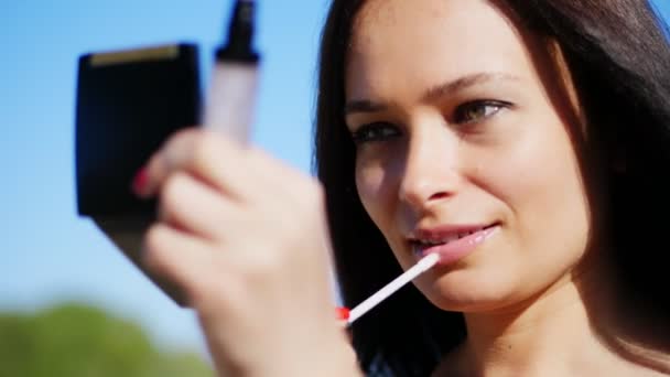 Portrait of a beautiful forty year old woman, brunette with green eyes. she looks in small mirror in powder box and colors lips with lip gloss. summer, against the blue sky — Stock Video