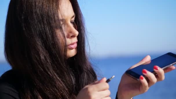 Estate, mattina. una bella quarantenne, dai lunghi capelli scuri, tiene in mano un cellulare, un dispositivo. prova a collegare la spina dalle cuffie al dispositivo . — Video Stock