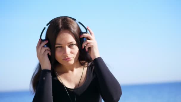 Verão, contra o fundo do mar. mulher bonita, com cabelo escuro longo, vestidos grandes fones de ouvido, escuta música ao ar livre, danças. mulher ouvindo música em fones de ouvido — Vídeo de Stock