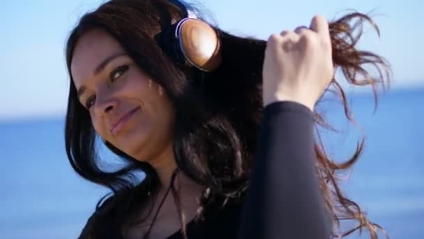 Summer, against the background of the sea. beautiful woman, with long dark hair, listening to music on headphones from a mobile phone, device, outdoors, dances. Enjoying Music — Stock Video