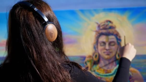 Verano, cerca del río, en la playa. al amanecer, una hermosa artista con grandes auriculares marrones sostiene un pincel en su mano, pinta en lienzo al dios Shiva. escuchar música, relajarse — Vídeo de stock