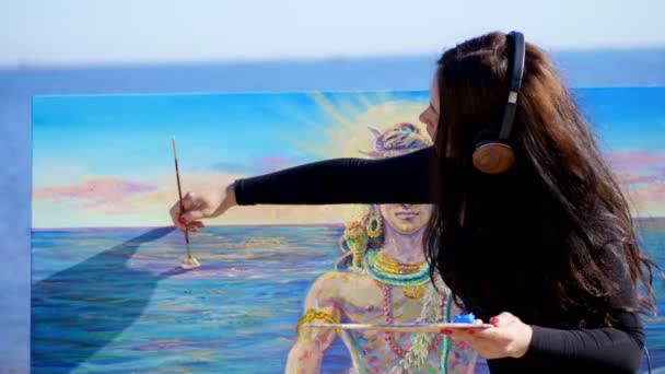 Verano, cerca del río, en la playa. al amanecer, una hermosa artista con grandes auriculares marrones sostiene un pincel, paleta en sus manos, pinturas sobre lienzo dios Shiva. escuchar música, relajarse — Vídeo de stock