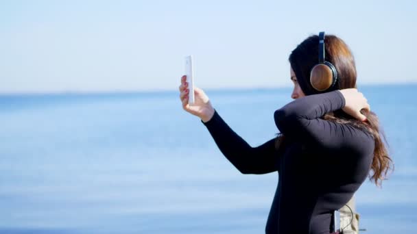 Sommaren, nära floden, på stranden. vid soluppgången, vacker kvinna, med långt mörkt hår, enhet, skjuta video av telefonen, lyssna på musik i hörlurar från en mobiltelefon, och gör selfi. — Stockvideo