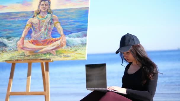 Καλοκαίρι, παραλία της θάλασσας. Ανατολή του ηλίου, όμορφη γυναίκα καλλιτέχνης στον ελεύθερο χρόνο από σχέδιο, εργάζονται σε φορητό υπολογιστή, επικοινωνία σε κοινωνικά δίκτυα, δίπλα της υπάρχει καβαλέτο με τον Θεό Σίβα εικόνα — Αρχείο Βίντεο