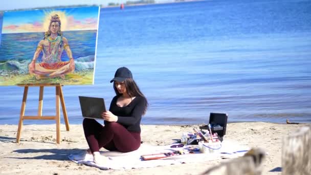 Sommer, Strand am Meer. Sonnenaufgang, schöne Künstlerin in ihrer Freizeit vom Zeichnen, Arbeiten am Laptop, Kommunikation in sozialen Netzwerken, daneben Staffelei mit Gott Shiva-Bild — Stockvideo