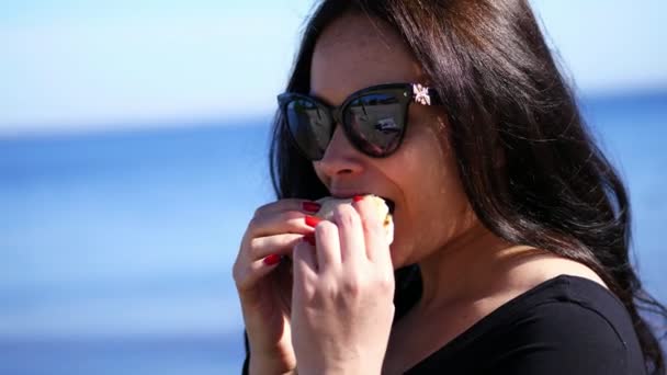 Léto, venku. portrét krásné bruneta žena, s dlouhými tmavými vlasy, nosí brýle, s apetite jíst fastfood, sexuálně kousání hamburger — Stock video
