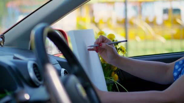 Kunstner, vakker brunette kvinne, sitter i bilen, tegner blyantskisse, tegner bukett med gule løvetann. trekke fra naturen, den kreative prosessen med å trekke. sommer – stockvideo