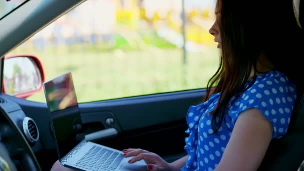 Όμορφη γυναίκα μελαχρινή σε ένα μπλε φόρεμα, κάθεται στο αυτοκίνητο, λειτουργεί σε φορητό υπολογιστή, επικοινωνία σε κοινωνικά δίκτυα. χρησιμοποιεί το διαδίκτυο, την κινητή επικοινωνία — Αρχείο Βίντεο