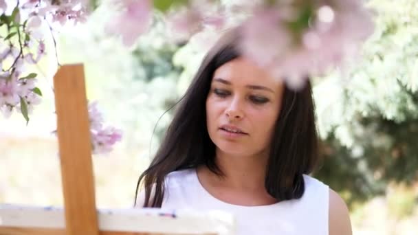 Portret van een mooie vrouw schilder in witte jurk, verven van de kunstenaar een beeld van de bloemen tijdens de bloei voorjaar appelboomgaard, ze geldt verf aan het canvas met een speciale kleine spatel, met behulp van een — Stockvideo